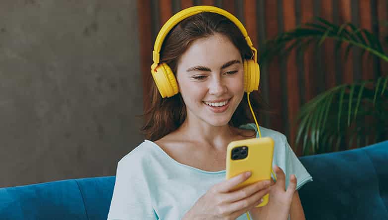 Une femme est assise sur le canapé en faisant défiler son téléphone et utilise en même temps son casque filaire.