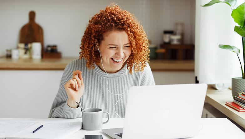 Une femme est assise devant son ordinateur portable, rit devant l'écran tout en portant des écouteurs avec fil.
