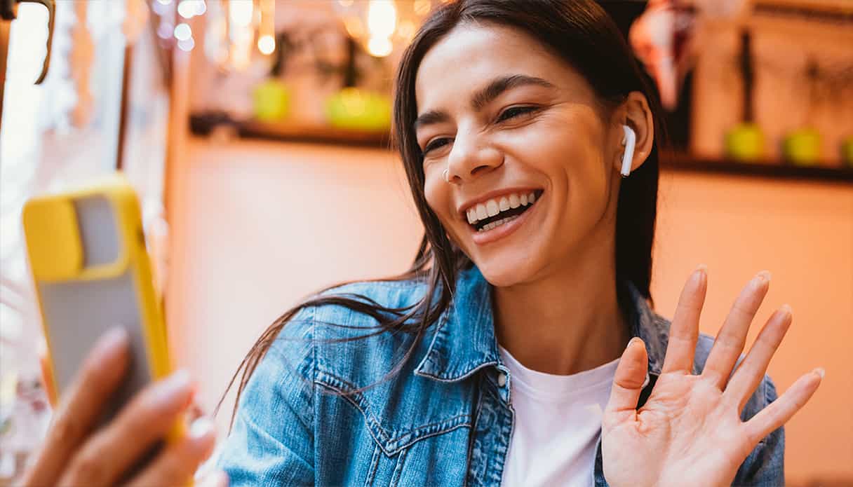 Une femme sourit en faisant signe à son téléphone, elle est en visioconférence et porte ses écouteurs sans fil.