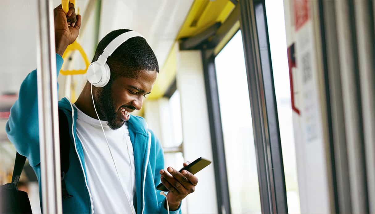 Un homme est debout dans le bus et regarde son téléphone en riant, il porte un casque avec fil.