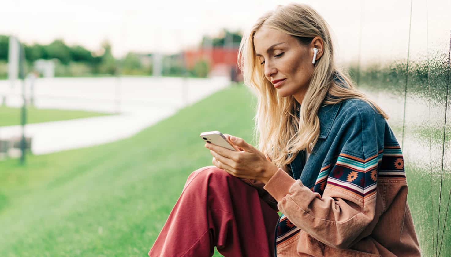 Une femme est assise sur une pelouse avec un téléphone dans ses mains, et elle a des écouteurs sans fil ses oreilles.